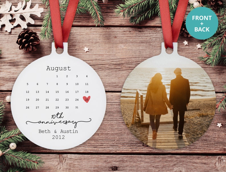 May include: White round ornament with a calendar for August with a red heart marking the 25th. The text "10th anniversary" and "Beth & Austin 2012" is below the calendar. The back of the ornament features a photo of a couple walking on a beach at sunset.