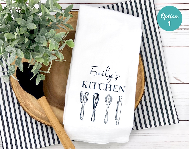 Puede incluir: Toalla de cocina blanca con el texto "Emily's Kitchen" y un gr&aacute;fico de cuatro utensilios de cocina: una esp&aacute;tula, un batidor, una cuchara y un rodillo.