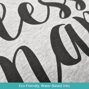 Puede incluir: Primer plano de una sudadera gris con la palabra "bless" impresa en negro, utilizando tintas ecol&oacute;gicas a base de agua.