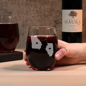 May include: Two stemless wine glasses with etched outlines of Georgia and Illinois, connected by a dotted line with hearts in each state. The glasses are filled with red wine.