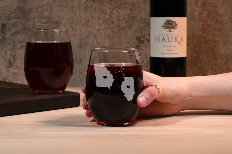 May include: Two stemless wine glasses with etched outlines of Georgia and Illinois, connected by a dotted line with hearts in each state. The glasses are filled with red wine.