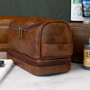 May include: A brown leather travel toiletry bag with two zippered compartments. The bag has a gold zipper and a brown leather handle.