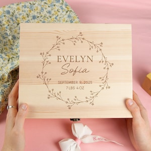 May include: A light-colored wooden keepsake box with the name "Evelyn Sofia" and birth details engraved within a floral wreath. The box is held by hands, with a floral fabric and a wooden block nearby.