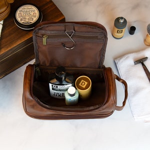 May include: A brown leather travel toiletry bag with a zipper closure and a handle. The bag is open and contains several bottles of toiletries, including a bottle of "TRUE FADE" hair product.