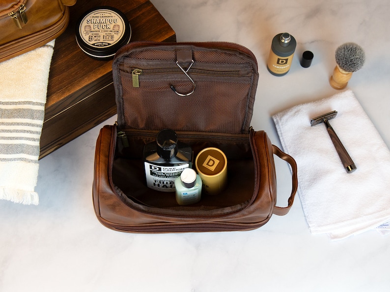May include: A brown leather travel toiletry bag with a zipper closure and a handle. The bag is open and contains several bottles of toiletries, including a bottle of "TRUE FADE" hair product.