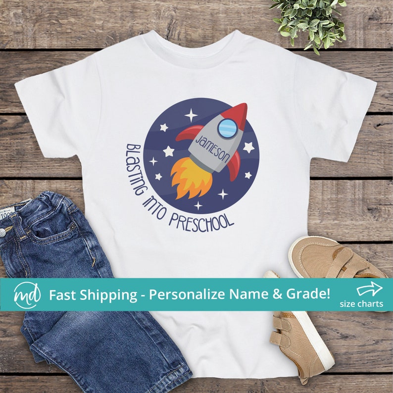 Puede incluir: Camiseta blanca con un gr&aacute;fico circular azul de un cohete con llamas y estrellas. El texto "Blastin' into Preschool" est&aacute; escrito alrededor del c&iacute;rculo. El nombre "Jameson" est&aacute; escrito en el cohete.