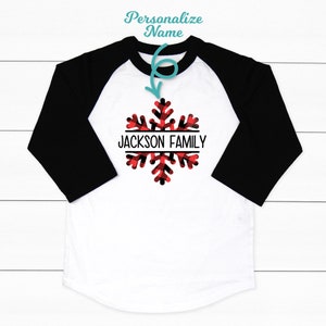 May include: White and black baseball tee shirt with red and black buffalo plaid snowflake design. The text "JACKSON FAMILY" is centered on the snowflake.