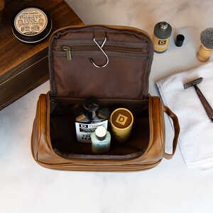 May include: A brown leather travel toiletry bag with a zipper closure and a hook. The bag is open and contains several bottles of toiletries.