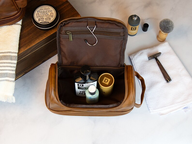 May include: A brown leather travel toiletry bag with a zipper closure and a hook. The bag is open and contains several bottles of toiletries.
