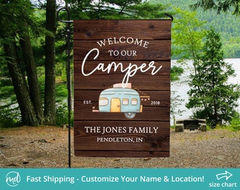 May include: A garden flag with a wooden plank design. The flag reads "WELCOME TO OUR CAMPER" in white script above a blue and yellow camper illustration. Below the camper is "THE JONES FAMILY, PENDLETON, IN". The flag is displayed outdoors.