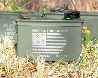 May include: An olive green metal ammunition box featuring a white distressed American flag graphic. The text reads PROPERTY OF: AUSTIN, GROOMSMAN, and 3.15.25. A small black firearm magazine with a visible bullet rests on the top lid of the container.