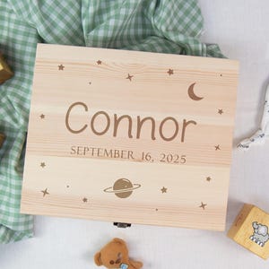 May include: A light-colored wooden keepsake box with the name "Connor" and the date "September 16, 2025" engraved on the lid. The lid is decorated with stars, a crescent moon, and a planet. Wooden blocks and a teddy bear are nearby.