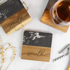 May include: A set of square coasters featuring a split design of black marble and natural wood. Each coaster is engraved with the name smith in a cursive script. One coaster is held in a gold metal holder, while another supports a glass of amber liquid.