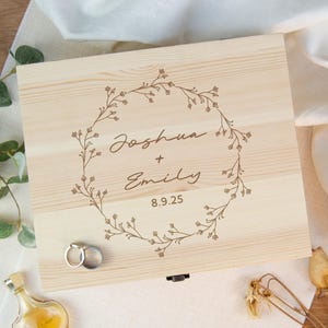 May include: A light-colored wooden box with a floral wreath design and the names "Joshua + Emily" and the date "8.9.25" engraved on the lid. Two silver rings are visible on the box.
