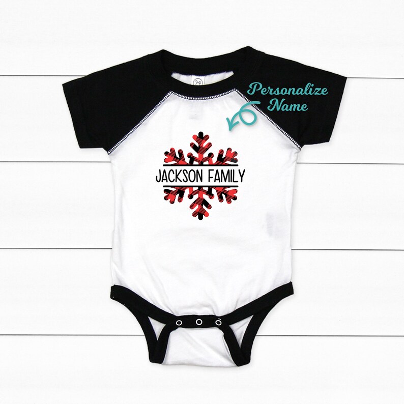 May include: White baby onesie with black raglan sleeves. The onesie features a red and black plaid snowflake with the text "JACKSON FAMILY" in a black banner.