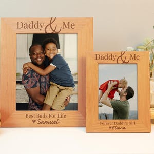 May include: Two wooden photo frames with the text "Daddy & Me" engraved on the top. The frames are both light brown and have a photo of a father and child in each. The left frame has the text "Best Buds For Life" and "Samuel" engraved below the photo. The right frame has the text "Forever Daddy's Girl" and "Eliana" engraved below the photo.