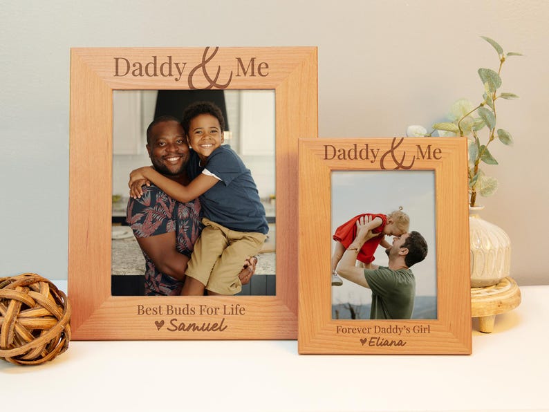 May include: Two wooden photo frames with the text "Daddy & Me" engraved on the top. The frames are both light brown and have a photo of a father and child in each. The left frame has the text "Best Buds For Life" and "Samuel" engraved below the photo. The right frame has the text "Forever Daddy's Girl" and "Eliana" engraved below the photo.