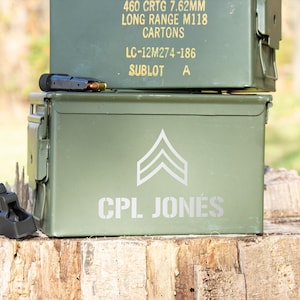 May include: A green metal ammo can with a silver chevron and the text "CPL JONES" printed on the front. The can is sitting on a wooden stump.