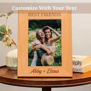 May include: A wooden picture frame with a light brown finish. The frame has a white mat and a clear glass front. The frame is decorated with the text "Best Friends" and "Abby + Elena". The frame contains a photo of two women smiling and hugging.