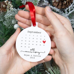 May include: White round ornament with a calendar for September with a red heart on the 13th. The ornament says "10th anniversary Bryan & Caitlin 2014".