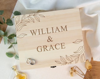 May include: A rectangular light-coloured wooden box with a hinged lid featuring the engraved text WILLIAM & GRACE in the centre. The lid is decorated with brown line art of leafy branches in the top-left and bottom-right corners. Two silver rings rest on the surface.