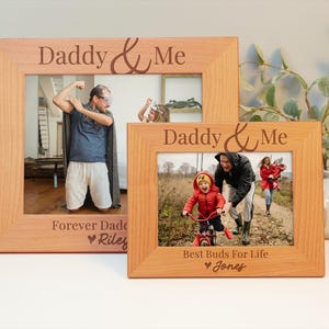 May include: Two wooden picture frames with the text "Daddy & Me" engraved on them. The larger frame has a photo of a man in a blue shirt and white shorts. The smaller frame has a photo of a father and son in a park. The larger frame has the text "Forever Dad Riley" engraved on the bottom. The smaller frame has the text "Best Buds For Life Jones" engraved on the bottom.