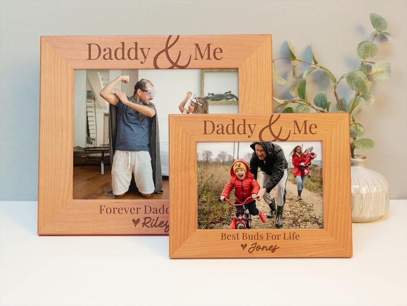 May include: Two wooden picture frames with the text "Daddy & Me" engraved on them. The larger frame has a photo of a man in a blue shirt and white shorts. The smaller frame has a photo of a father and son in a park. The larger frame has the text "Forever Dad Riley" engraved on the bottom. The smaller frame has the text "Best Buds For Life Jones" engraved on the bottom.