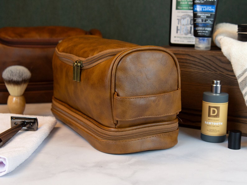 May include: A brown leather travel toiletry bag with two zippered compartments and a strap. The bag is sitting on a white surface.