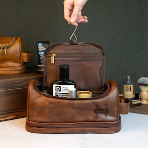 May include: Brown leather travel toiletry bag with a zipper closure and a personalized monogram "N NELSON". The bag is open and contains various toiletries, including a bottle of hair wash, a small tube of cream, and a small bottle of cologne.