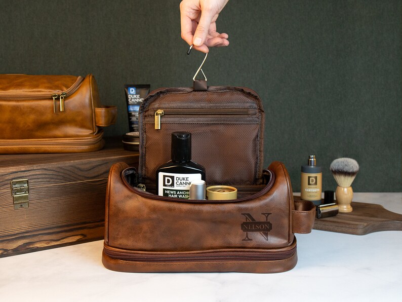 May include: Brown leather travel toiletry bag with a zipper closure and a personalized monogram "N NELSON". The bag is open and contains various toiletries, including a bottle of hair wash, a small tube of cream, and a small bottle of cologne.
