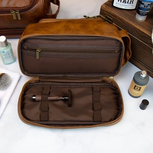 May include: A brown leather travel toiletry bag with a zippered closure. The bag is open and the interior is lined with brown fabric. The bag has multiple compartments and pockets for storing toiletries. A razor is visible in one of the compartments.