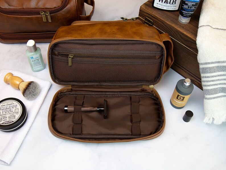 May include: A brown leather travel toiletry bag with a zippered closure. The bag is open and the interior is lined with brown fabric. The bag has multiple compartments and pockets for storing toiletries. A razor is visible in one of the compartments.
