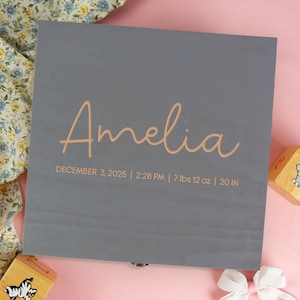May include: A square gray wooden keepsake box featuring the name Amelia in light orange script lettering. Below the name, the text reads DECEMBER 3, 2025 | 2:28 PM | 7 lbs 12 oz | 20 IN. The box has a smooth finish and a metal latch at the bottom edge.