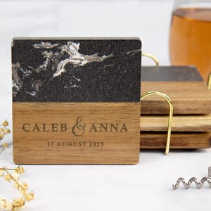 May include: A set of square coasters featuring a combination of dark black marble with white veining and natural brown wood. The wooden section is engraved with the text CALEB & ANNA and the date 17 AUGUST 2025. The coasters are held in a gold metal stand.