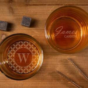 May include: Two clear glass whiskey glasses with a geometric pattern and a letter W on one glass and the name "James Carson" on the other glass. The glasses are on a wooden surface with two black square ice cubes.