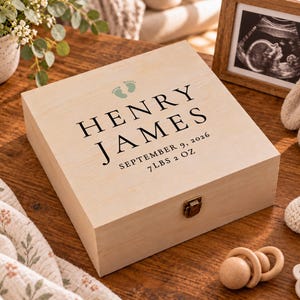 May include: A square light-colored wooden keepsake box with a metal latch. The lid features the printed text "HENRY JAMES", "SEPTEMBER 9, 2026", and "7 LBS 2 OZ" in black, accompanied by two small green baby footprint icons centered above the name.