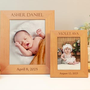 May include: Two wooden picture frames with engraved names and dates. The left frame has a photo of a baby sleeping and the right frame has a photo of a toddler girl sitting in a chair. The left frame says "Asher Daniel April 8, 2025" and the right frame says "Violet Ava August 12, 2025".