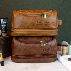 May include: Two brown leather travel toiletry bags with zipper closures. The top bag has the text "MALEY designs Personalized Clothing & Gifts" embossed on the front. The bottom bag has the initials "md" embossed on the front.