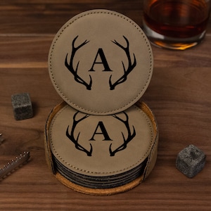 May include: Set of six brown leather coasters with a black antler design and the letter "A" in the center. The coasters are stacked in a circular holder.