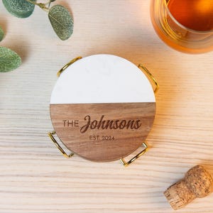 May include: A circular coaster featuring a split design with a white top half and a brown wood bottom half. The wood section is engraved with the text THE Johnsons EST. 2024. The coaster rests in a gold-toned metal holder with four curved wire legs.