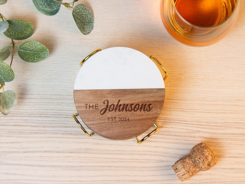 May include: A circular coaster featuring a split design with a white top half and a brown wood bottom half. The wood section is engraved with the text THE Johnsons EST. 2024. The coaster rests in a gold-toned metal holder with four curved wire legs.