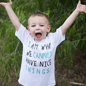 Puede incluir: Un ni&ntilde;o peque&ntilde;o con una camiseta blanca con el texto "I am why we cannot have nice things." en letras negras y verde azulado. Lleva pantalones vaqueros azules y tiene los brazos levantados en el aire.
