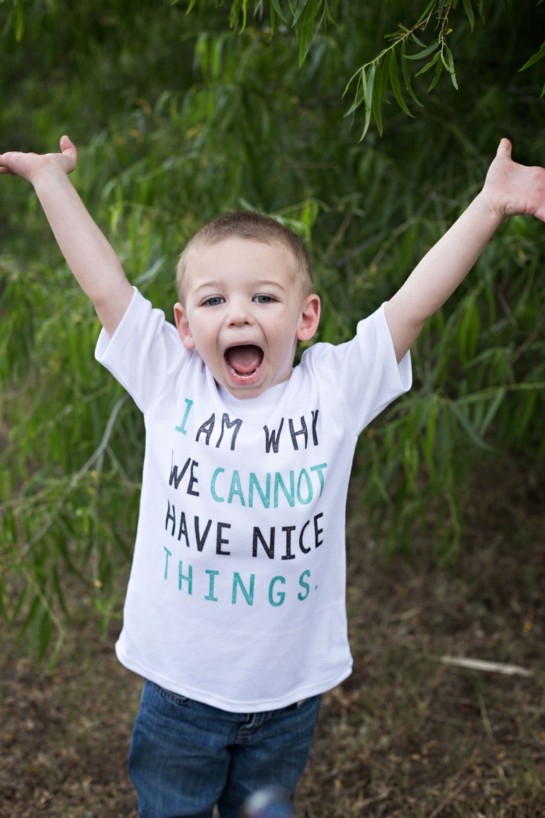 Puede incluir: Un ni&ntilde;o peque&ntilde;o con una camiseta blanca con el texto "I am why we cannot have nice things." en letras negras y verde azulado. Lleva pantalones vaqueros azules y tiene los brazos levantados en el aire.
