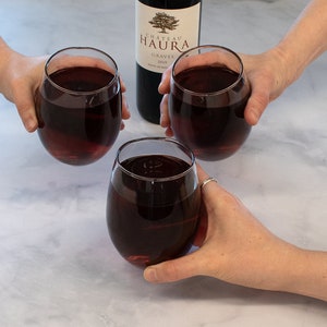 May include: Three glasses of red wine are held up in a toast. The wine is a deep red color and the glasses are clear. A bottle of Chateau Haura Graves wine is in the background.
