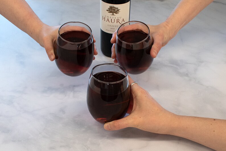 May include: Three glasses of red wine are held up in a toast. The wine is a deep red color and the glasses are clear. A bottle of Chateau Haura Graves wine is in the background.