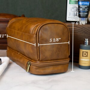 May include: Brown leather travel toiletry bag with two zippered compartments. The bag measures 11 inches wide, 6 1/4 inches tall, and 5 1/8 inches deep.
