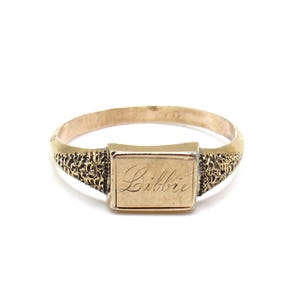 Könnte beinhalten: Goldfarbener Ring mit rechteckiger Platte, auf der der Name "Libbie" in Schreibschrift eingraviert ist. Das Band weist auf beiden Seiten der Platte ein detailliertes, strukturiertes Design auf. Der Ring ist vor weißem Hintergrund platziert.