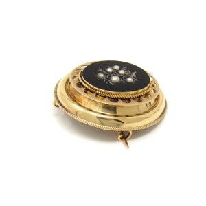 May include: A gold brooch with a black enamel oval center featuring a white pearl floral design. The brooch has a decorative, circular, openwork frame.