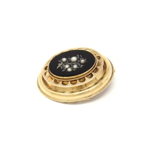 May include: A gold oval brooch with a black enamel center featuring a floral design with pearl accents.