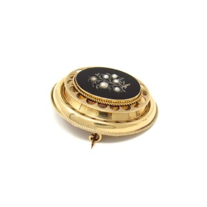 May include: A gold brooch with a black enamel center featuring a pearl-encrusted floral design. The brooch has a decorative, scalloped edge.